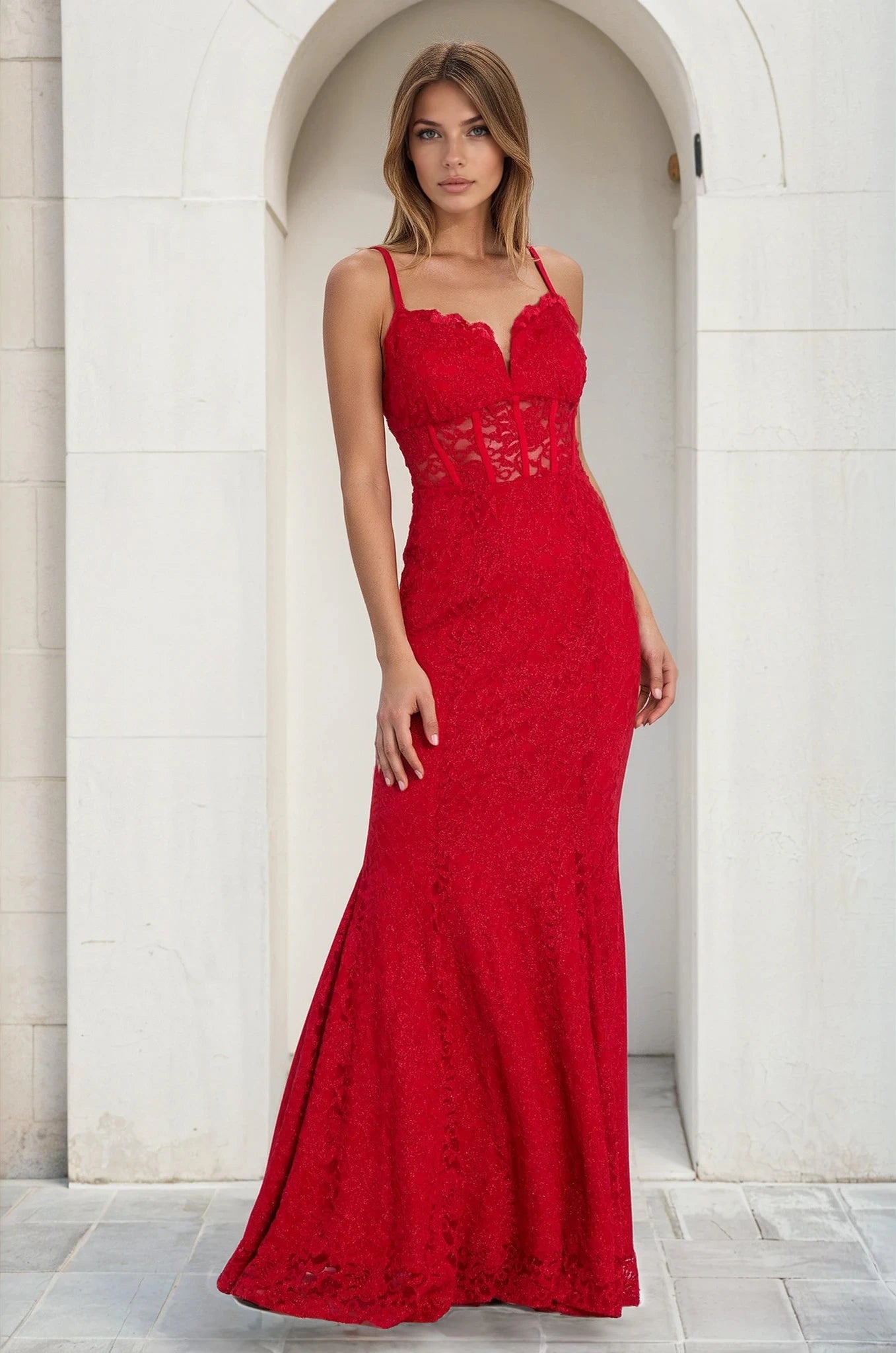 Woman wears the Long Red Lace Prom Dress: Morgan 19069—a fitted, floor-length mermaid gown with thin straps and a semi-sheer bodice—while standing in front of a white arched doorway.